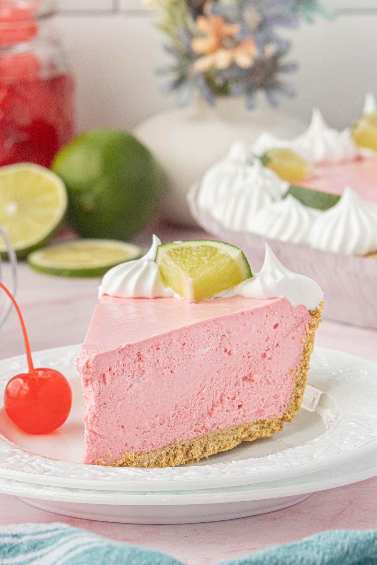 Slice of No-Bake Cherry Limeade Pie garnished with whipped cream and a fresh lime wedge on a white dessert plate with cherries nearby.