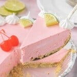 Slice of No-Bake Cherry Limeade Pie balanced on a dessert fork with whipped cream, lime wedges, and the whole pie blurred in the background.