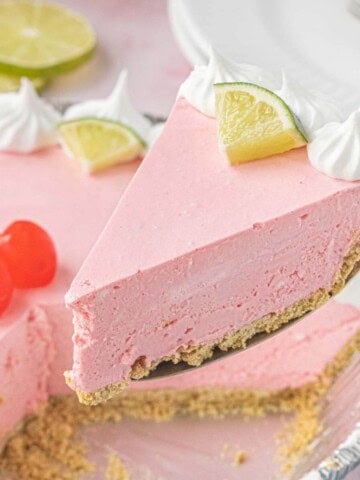 Slice of No-Bake Cherry Limeade Pie balanced on a dessert fork with whipped cream, lime wedges, and the whole pie blurred in the background.