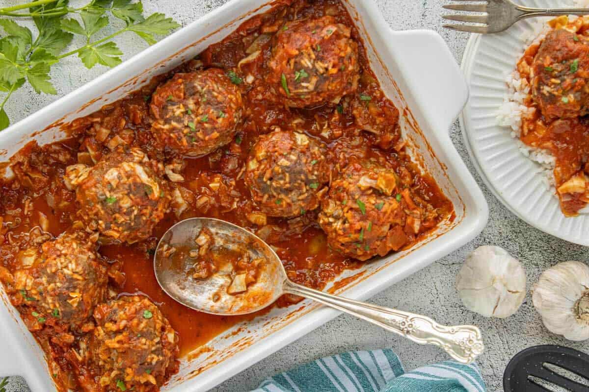 Baked porcupine meatballs in a casserole dish with tomato sauce, cabbage, and fresh parsley. Several meatballs have been removed and are served on a plate over rice.