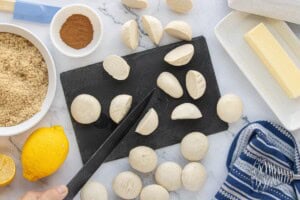 Cut frozen dinner roll dough into halves on a cutting board with ingredients arranged nearby.