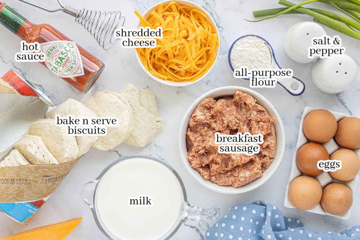 Ingredients for biscuits and gravy casserole including breakfast sausage, biscuit dough, eggs, milk, shredded cheese, flour, hot sauce, salt, and pepper arranged on a countertop.