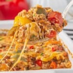 Serving spoon lifting a cheesy bite of casserole filled with ground beef, rice, and colorful bell peppers, with melted cheese stretching from the baking dish below.