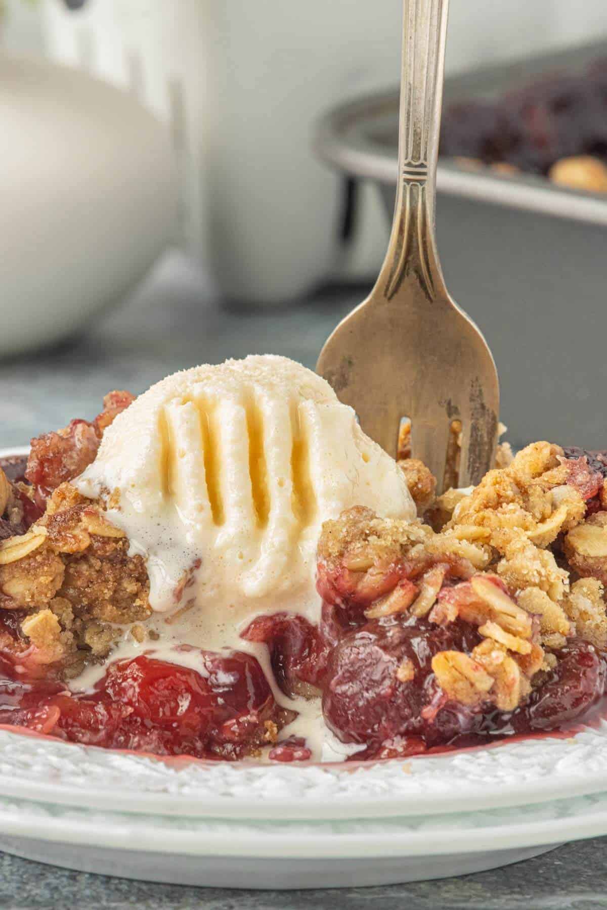 Individual serving of cherry crisp topped with melted vanilla ice cream on a dessert plate with a fork ready to take another bite.