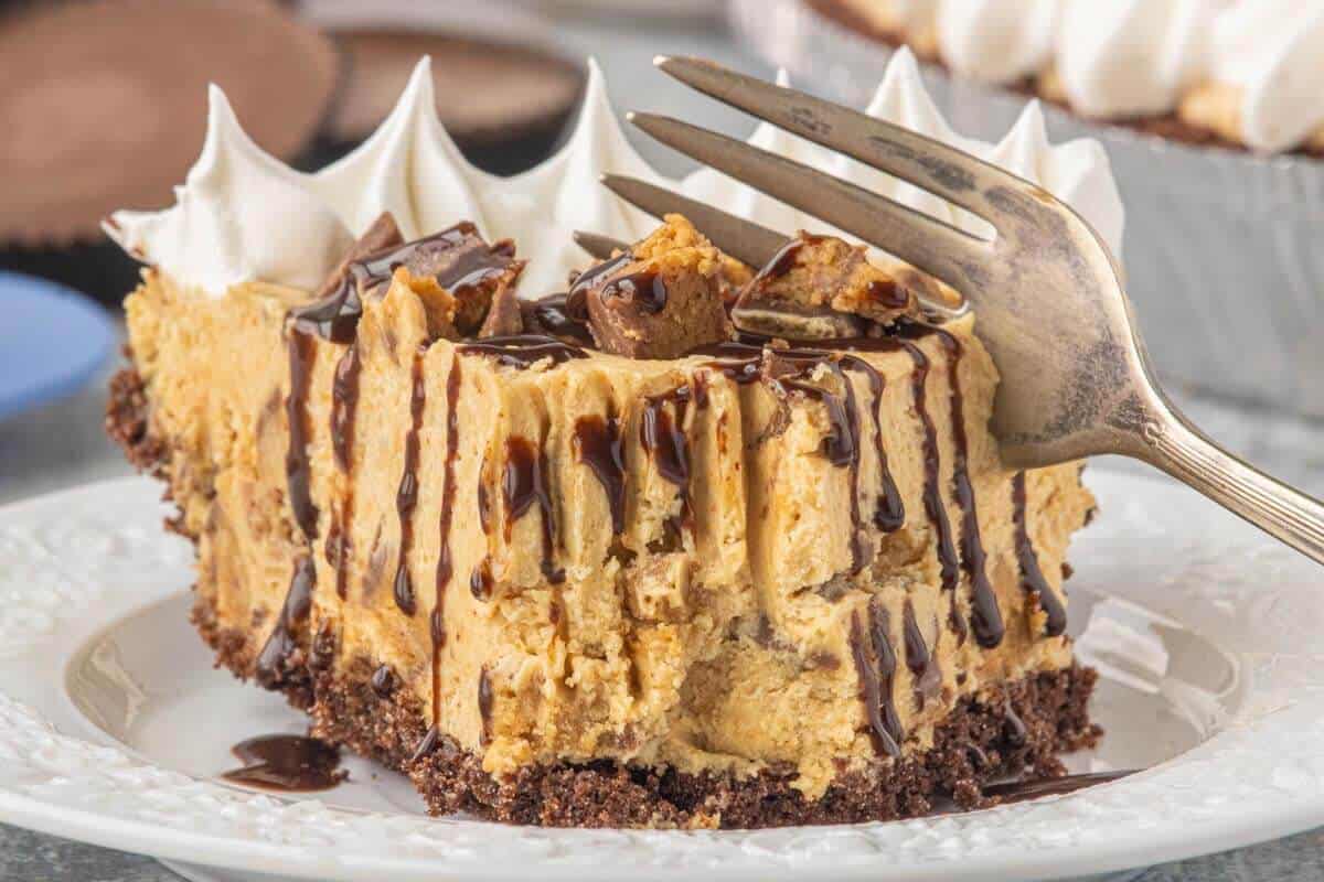 Close-up of a slice of no-bake peanut butter cup cheesecake with a chocolate cookie crust, peanut butter cup pieces, and drizzled with hot fudge.