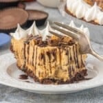 A slice of peanut butter chocolate cheesecake drizzled with hot fudge on a white plate, with the remaining pie in the background.