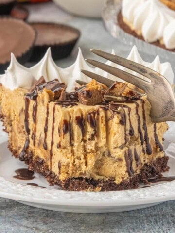 A slice of peanut butter chocolate cheesecake drizzled with hot fudge on a white plate, with the remaining pie in the background.