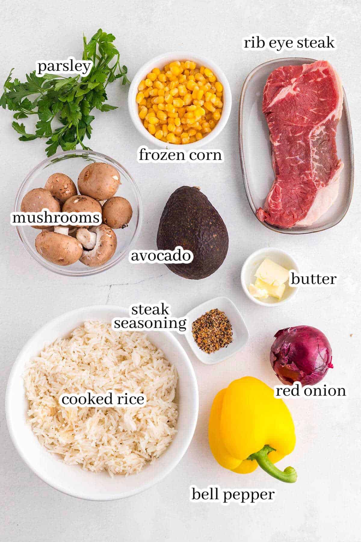ingredients on white counter top to make steak bowl including cooked rice, bell pepper, seasoning, avocado, red onion, butter, mushrooms, parsley, corn and steak.