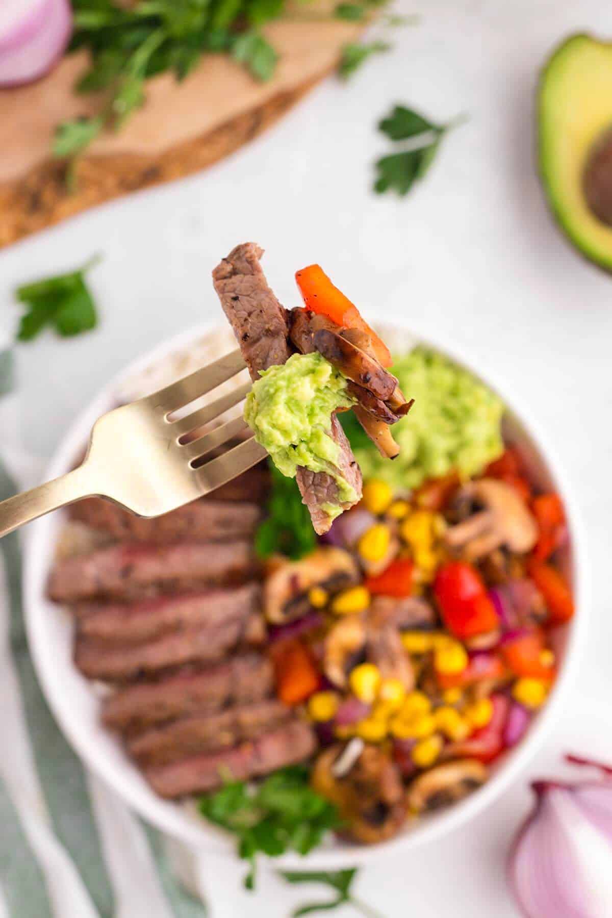 Close-up of a forkful of steak, mushrooms, peppers, and avocado from a homemade steak rice bowl recipe.