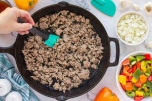 Ground beef browning in a cast iron skillet with diced onions, bell peppers, and garlic nearby.