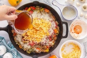 Beef broth being poured into a skillet filled with seasoned ground beef, rice, tomatoes, and shredded cheese.