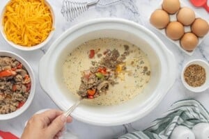 Cooked sausage and vegetables being added to the egg mixture in a slow cooker.