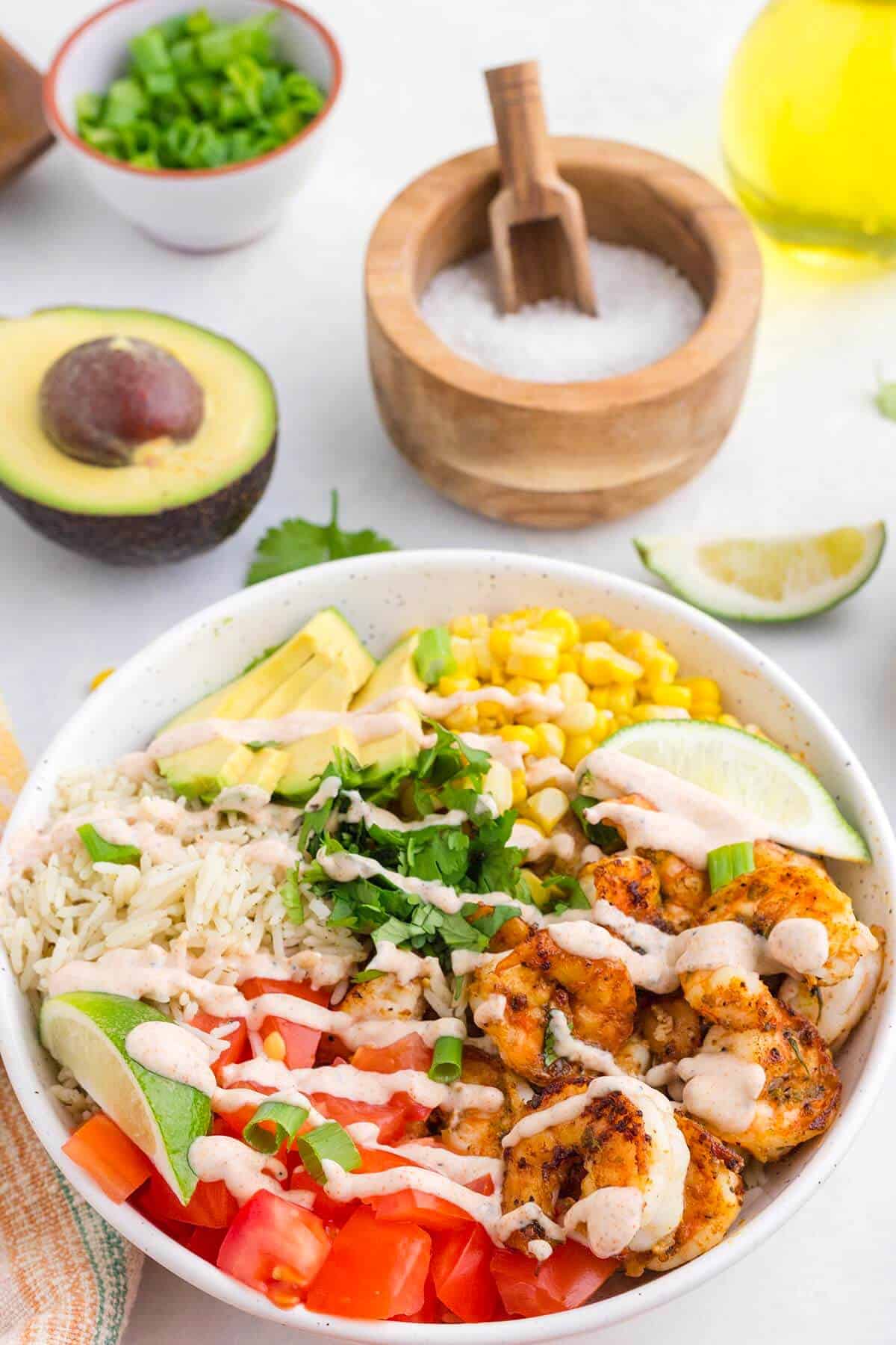 Overhead view of a homemade shrimp burrito bowl with rice, seasoned shrimp, avocado, tomatoes, corn, cilantro, lime wedges, and creamy lime dressing.