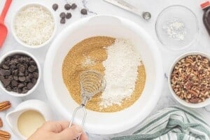 Hand whisk combining graham cracker crumbs, flour, powdered sugar, and baking powder in a large white mixing bowl surrounded by chocolate chips, coconut, chopped pecans, and sweetened condensed milk.