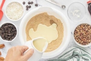 Sweetened condensed milk being poured into a bowl of dry ingredients before mixing with chocolate chips, coconut, and chopped pecans.