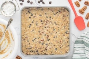 Prepared batter spread evenly into a square baking pan, ready to bake, with baking ingredients arranged around the pan on a marble countertop.