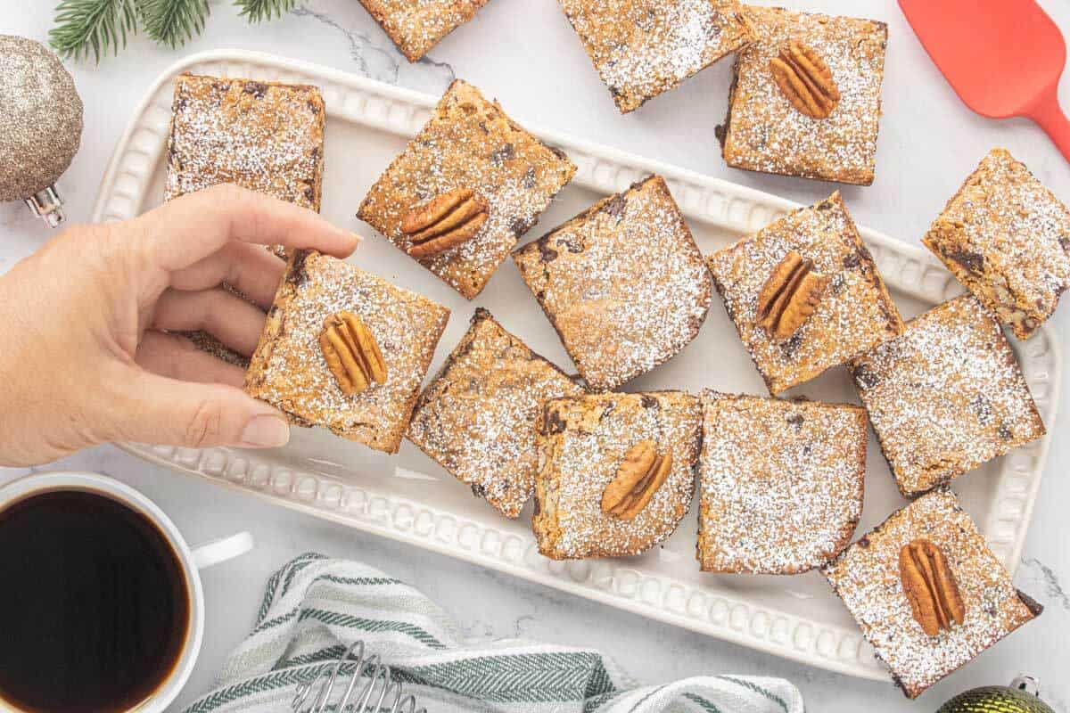Hand reaching for a powdered sugar dusted Graham Cracker Pecan Cookie Bar from a holiday serving platter with coffee and Christmas decorations.