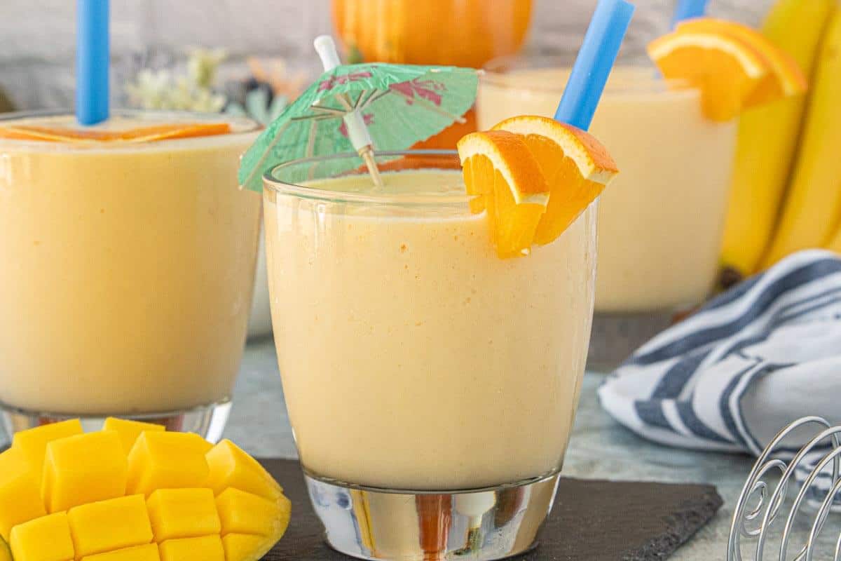 Three glasses of Mango Orange Creamsicle Smoothies served with fresh orange slices and diced mango on the side.