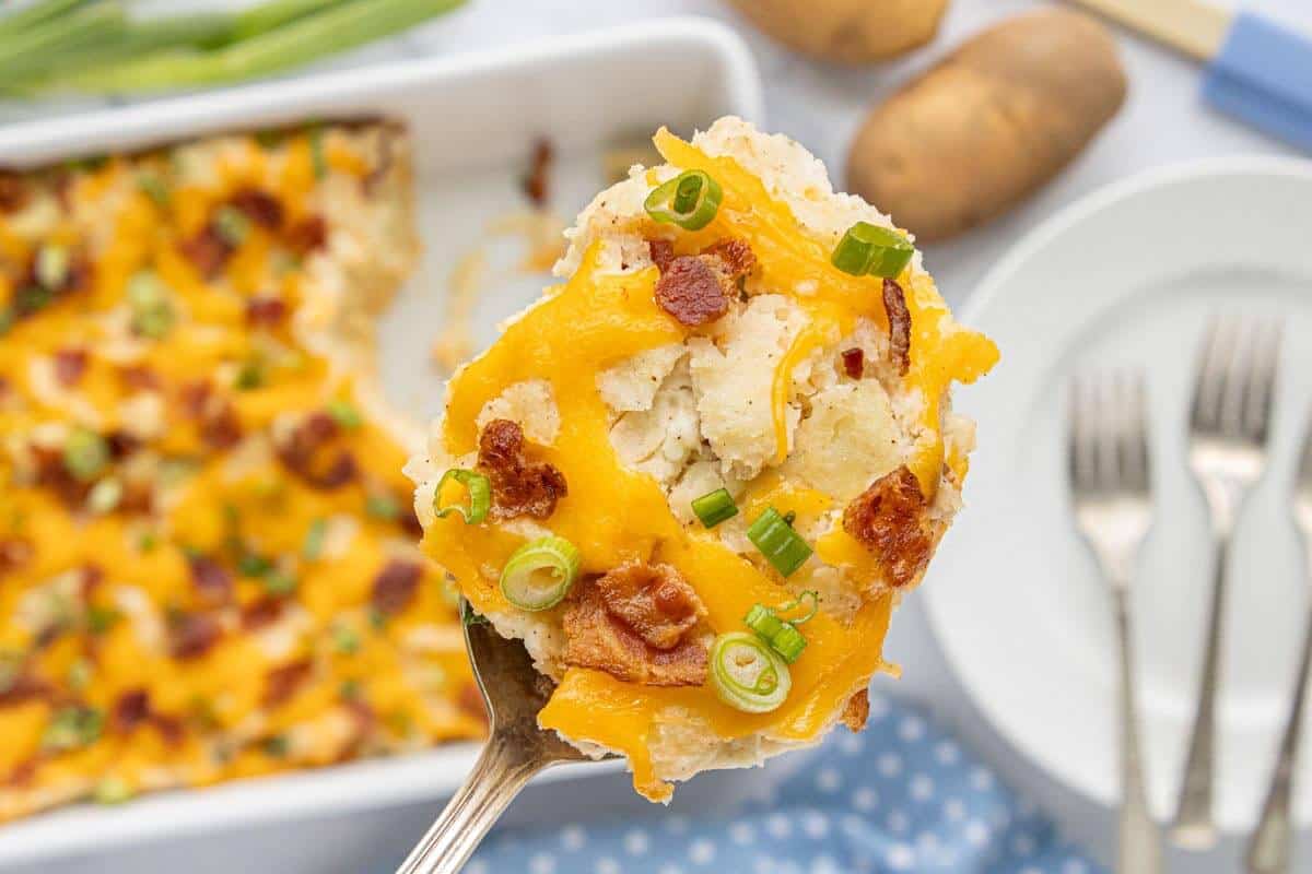 Spoon holding a bite of Million Dollar Potato Casserole with melted cheddar cheese, bacon, and green onions in front of the baking dish.