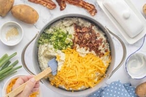 Shredded cheddar cheese, crispy bacon, and sliced green onions being folded into mashed potatoes.