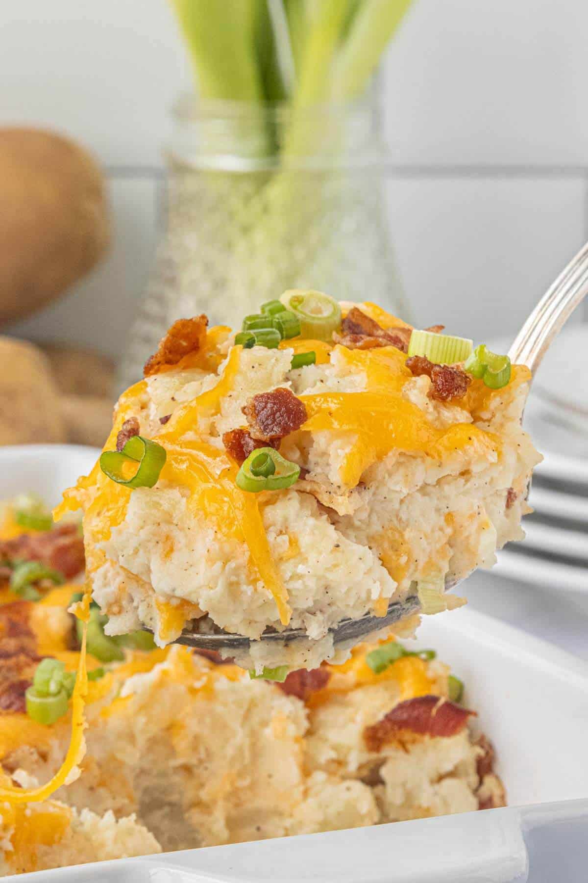 A serving of Million Dollar Potato Casserole with melted cheddar cheese, bacon, and green onions on a serving spoon.