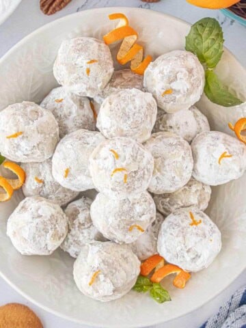 An overhead shot of a serving bowl filled with Orange Snowballs dusted with powdered sugar and garnished with fresh orange zest and mint leaves.