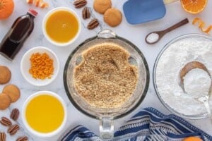 Crushed vanilla wafers and pecans in a mixing bowl, surrounded by butter, powdered sugar, salt, orange zest and oj concentrate.