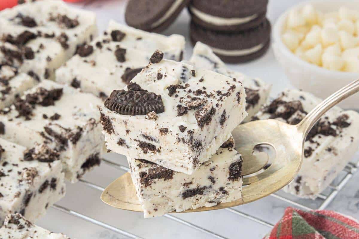 Two squares of homemade Oreo fudge on a serving spoon with more fudge pieces cooling on a wire rack in the background.