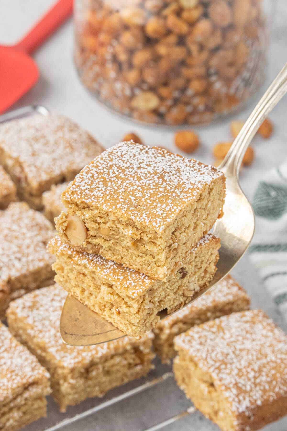 A spatula holds two peanut butter blondies dusted with powdered sugar above a cooling rack filled with freshly baked blondie bars.