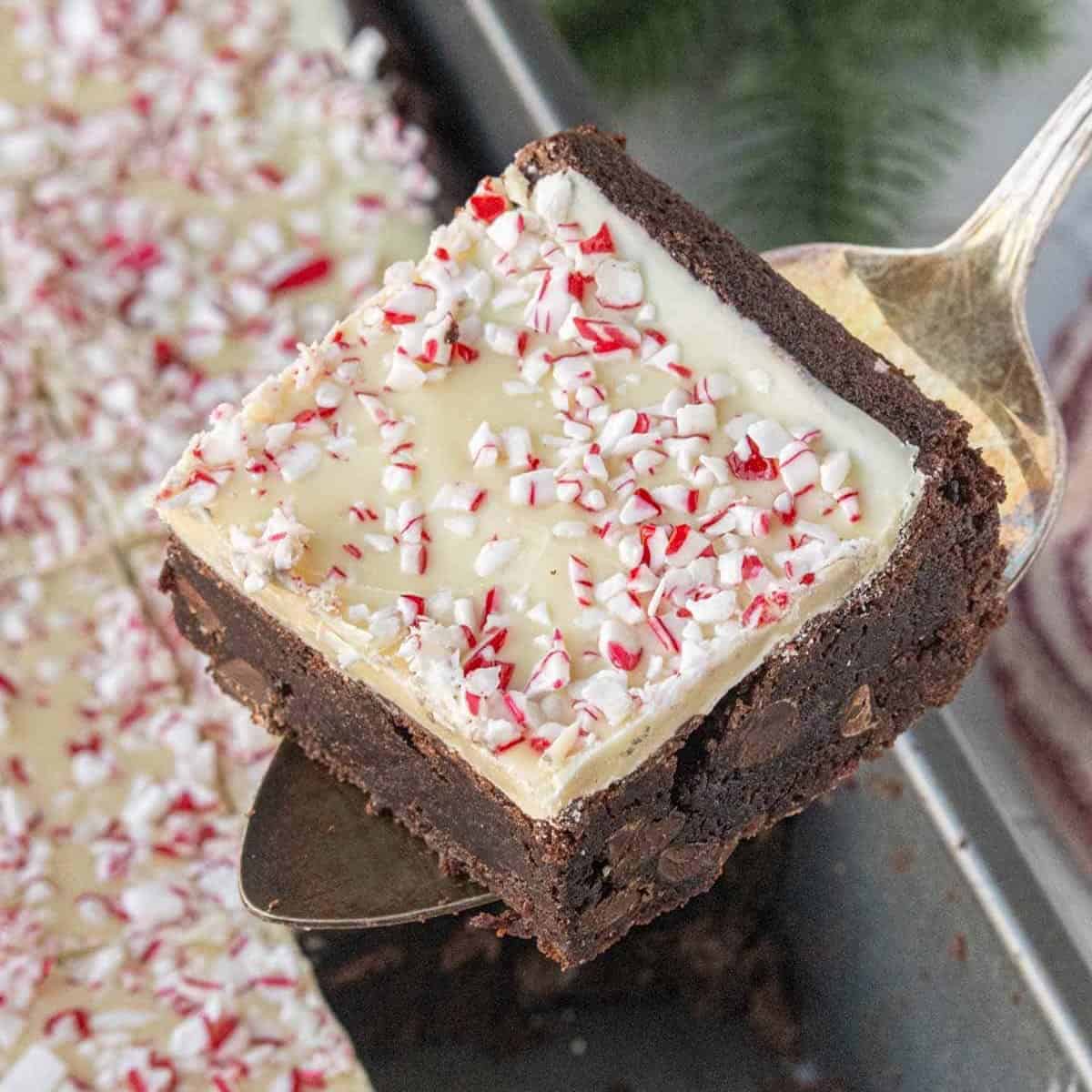 Close-up of a fudgy peppermint bark brownie topped with a layer of white chocolate and crushed peppermint candy, being removed from the baking sheet.