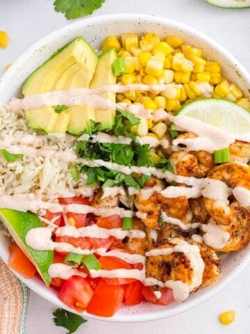 Shrimp burrito bowl with seasoned shrimp, rice, avocado, tomatoes, corn, cilantro, and creamy lime dressing in a white bowl.