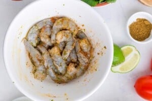 Raw shrimp tossed with olive oil, lime juice, lime zest, cumin, and chili powder in a white mixing bowl before cooking.