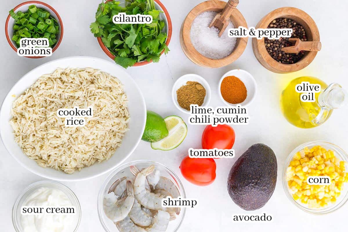 Overhead view of the ingredients for a shrimp burrito bowl, including cooked rice, raw shrimp, avocado, corn, tomatoes, cilantro, green onions, lime, sour cream, olive oil, cumin, chili powder, salt, and black pepper, with each ingredient labeled.