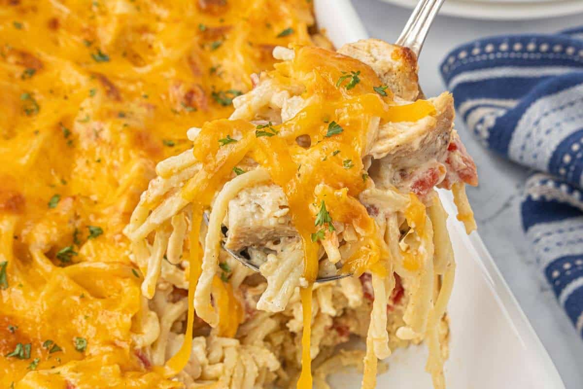 Spoon lifting a cheesy serving of chicken spaghetti from a white baking dish, topped with melted cheddar cheese and parsley.