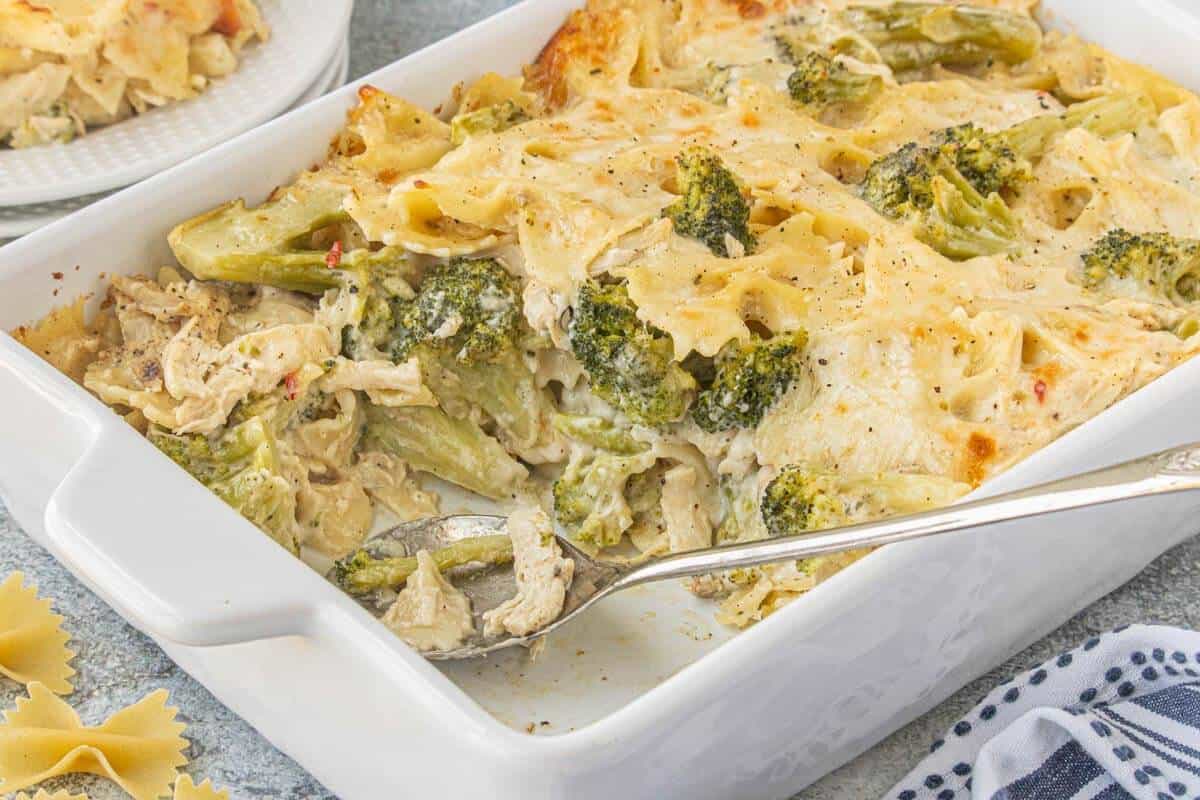 A casserole dish filled with cheesy chicken broccoli pasta casserole. Several servings have been removed. A spoon is resting in the bowl.