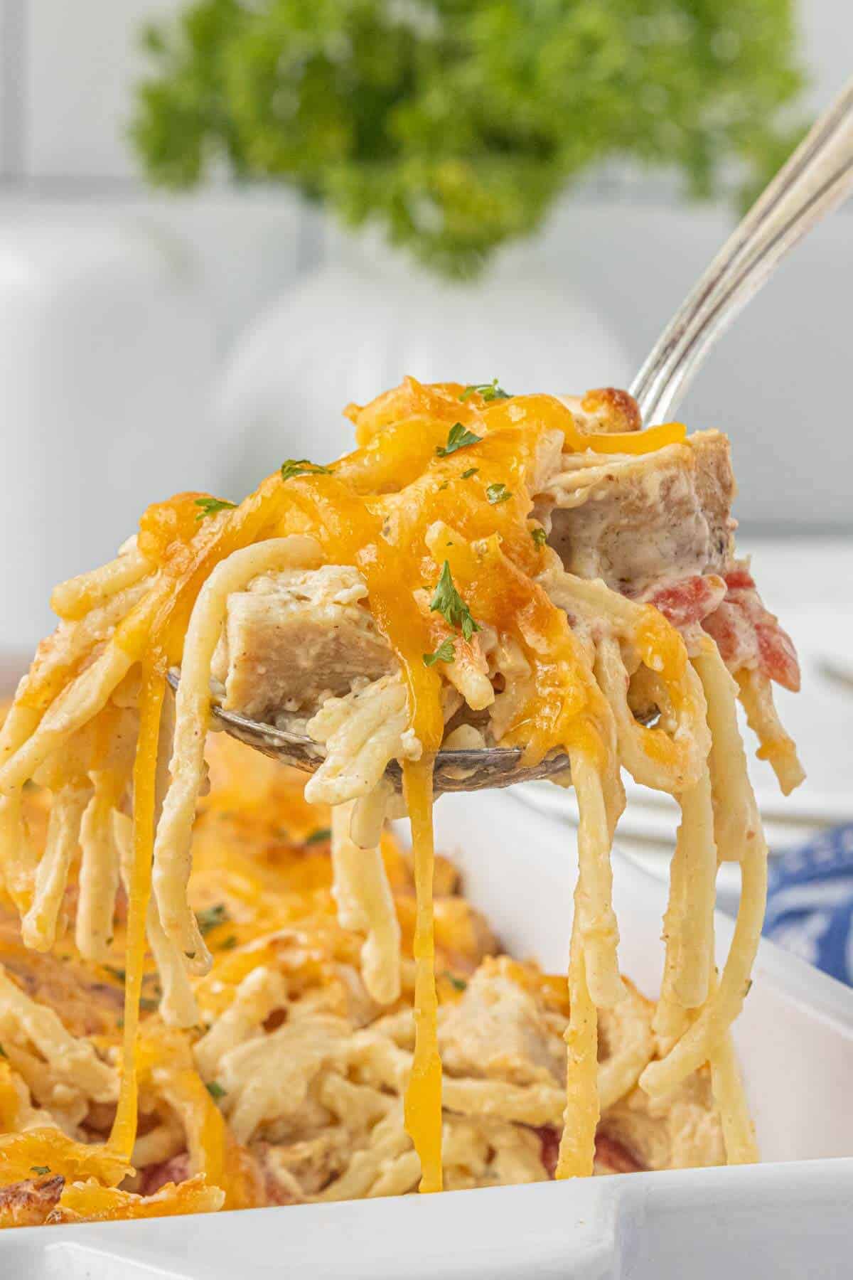 Spoon lifting a cheesy serving of chicken spaghetti with Rotel tomatoes from a white baking dish, topped with melted cheddar cheese and parsley.