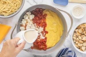 Cream cheese, condensed cheddar soup, Rotel tomatoes, chicken broth, and milk being added to a large skillet to make the creamy sauce for the casserole dish.