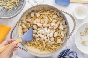 Cooked spaghetti and diced chicken being stirred into the creamy Rotel cheese sauce in a large skillet with a spatula.