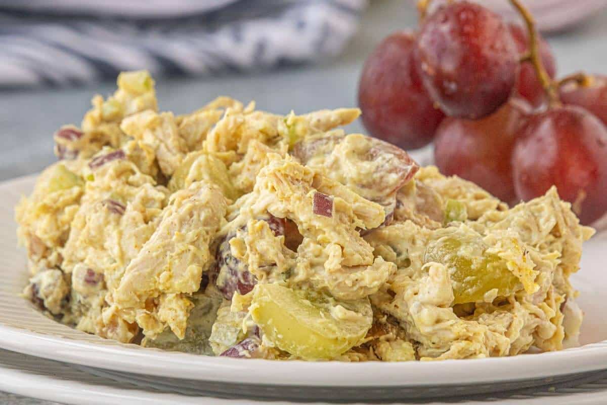 Curry chicken salad with shredded chicken, red and green grapes, celery, and red onion served on a white plate with a bunch of grapes.