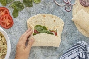 Hands folding a flour tortilla around curry chicken salad, tomatoes, red onion, and fresh spinach to make a wrap.