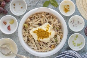 Mayonnaise and yellow curry powder added to shredded chicken, grapes, celery, red onion, and walnuts in a mixing bowl.