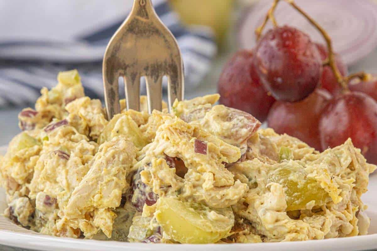 Creamy curry chicken salad with red and green grapes, celery, red onion, and shredded chicken served on a white plate. A fork is in the salad, ready to grab a bite.