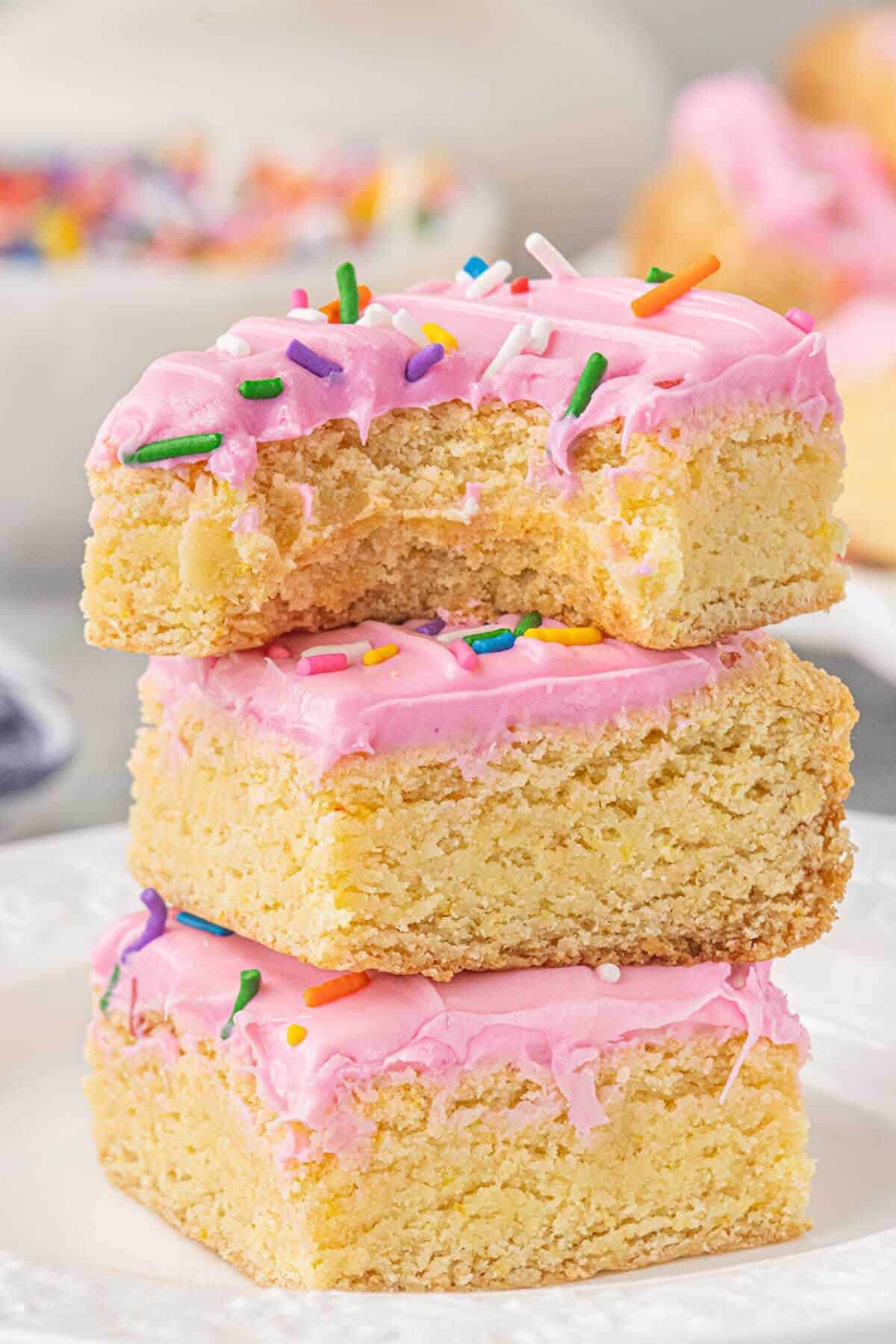Stack of three Lofthouse sugar cookie bars frosted with pink buttercream and rainbow sprinkles, with a bite taken from the top bar.