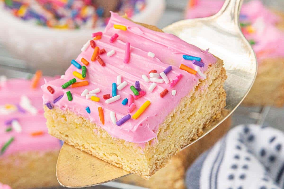Frosted sugar cookie bar topped with pink buttercream frosting and rainbow jimmies served on a metal spatula.