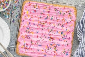 Cake mix Lofthouse sugar cookie bars topped with pink buttercream frosting and rainbow jimmies before slicing into squares.