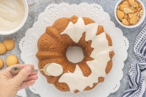 Vanilla glaze is spooned over a cooled Vanilla Wafer Bundt Cake on a white cake stand before topping with crushed vanilla wafers.