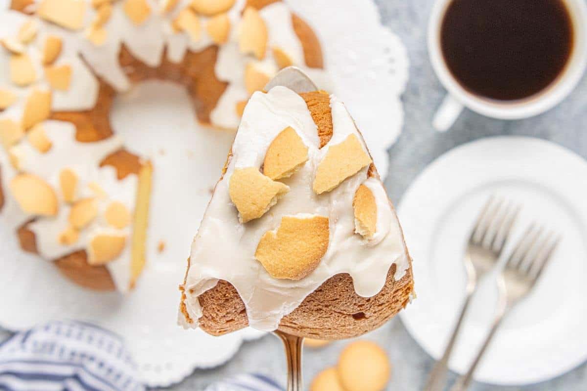 A slice of Vanilla Wafer Bundt Cake topped with vanilla glaze and crushed vanilla wafers is held on a cake server with the Bundt cake in the background.