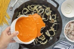 Jarred vodka sauce being poured into a skillet with sautéed onions.
