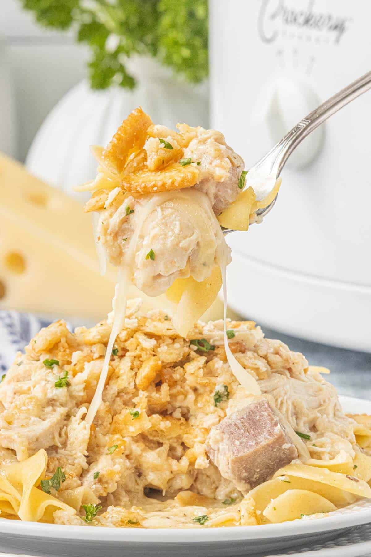 Forkful of creamy chicken, ham, melted Swiss cheese, and buttery cracker topping lifted from a plate of egg noodles.