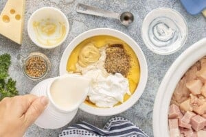 Evaporated milk being poured into a bowl with cream of chicken soup, sour cream, Dijon mustard, Worcestershire sauce, and Montreal steak seasoning.